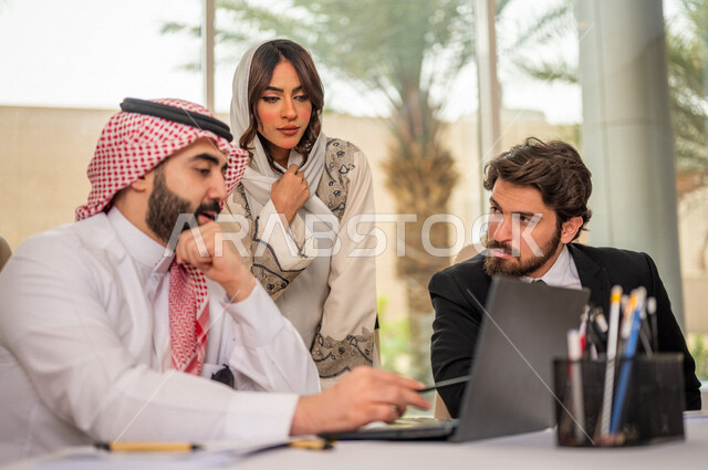 رجل اعمال عربي سعودي خليجي بالثوب السعودي التقليدي في اجتماع عمل مع عميل جديد يرتدي البدلة الرسمية، سيدة اعمال عربية سعودية خليجية تعمل على دراسة وتحليل المشاريع الجديدة، استخدام الحاسوب المحمول في تطوير العمل، اجتماعات عمل، الشراكة والتعاون