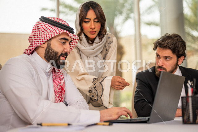 A Saudi Arabian Gulf businessman in the traditional Saudi dress in a business meeting with a new client wearing a formal suit, a Saudi Arabian Gulf business woman working on the study and analysis of new projects, the use of a laptop in business developme