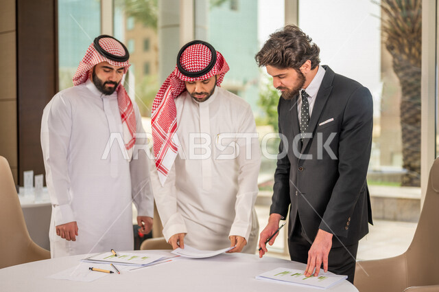 رجل اعمال عربي سعودي خليجي بالثوب السعودي التقليدي يقوم بتوقيع اتفاقية عمل لمشروع جديد مع عميل يرتدي البدلة الرسمية، رجل اعمال خليجي يشارك في نجاح صفقات العمل، التحقق والتدقيق في شروط العقد، قراءة البنود الخاصة بالعقد، الموافقة على الشروط، توقيع العقود