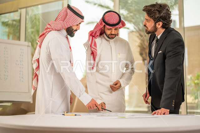 رجل اعمال عربي سعودي خليجي بالثوب السعودي التقليدي يقوم بتوقيع اتفاقية عمل لمشروع جديد مع عميل يرتدي البدلة الرسمية، رجل اعمال خليجي يشارك في نجاح صفقات العمل، التحقق والتدقيق في شروط العقد، قراءة البنود الخاصة بالعقد، الموافقة على الشروط، توقيع العقود