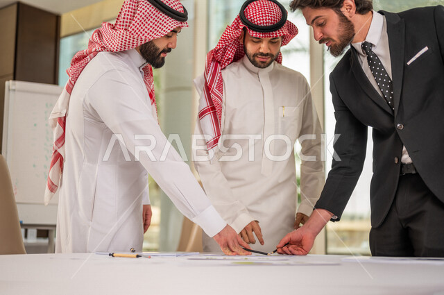 رجل اعمال عربي سعودي خليجي بالثوب السعودي التقليدي يقوم بتوقيع اتفاقية عمل لمشروع جديد مع عميل يرتدي البدلة الرسمية، رجل اعمال خليجي يشارك في نجاح صفقات العمل، التحقق والتدقيق في شروط العقد، قراءة البنود الخاصة بالعقد، الموافقة على الشروط، توقيع العقود