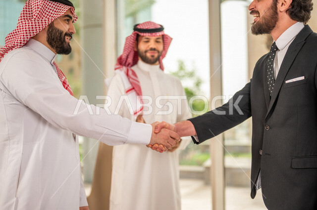 رجل اعمال عربي سعودي خليجي بالثوب السعودي التقليدي يتصافح مع عميل جديد يرتدي البدلة الرسمية، توقيع اتفاقية عمل لمشروع جديد، اتفاق عمل بين رجال الاعمال، صفقة ناجحة، تعاون تجاري، الوحدة والعمل الجماعي، الموافقة على شروط العمل، توقيع عقود العمل  