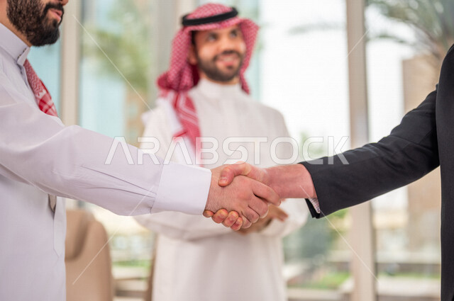 رجل اعمال عربي سعودي خليجي بالثوب السعودي التقليدي يتصافح مع عميل جديد يرتدي البدلة الرسمية، توقيع اتفاقية عمل لمشروع جديد، اتفاق عمل بين رجال الاعمال، صفقة ناجحة، تعاون تجاري، الوحدة والعمل الجماعي، الموافقة على شروط العمل، توقيع عقود العمل  