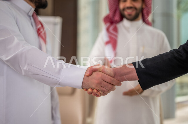 رجل اعمال عربي سعودي خليجي بالثوب السعودي التقليدي يتصافح مع عميل جديد يرتدي البدلة الرسمية، توقيع اتفاقية عمل لمشروع جديد، اتفاق عمل بين رجال الاعمال، صفقة ناجحة، تعاون تجاري، الوحدة والعمل الجماعي، الموافقة على شروط العمل، توقيع عقود العمل  