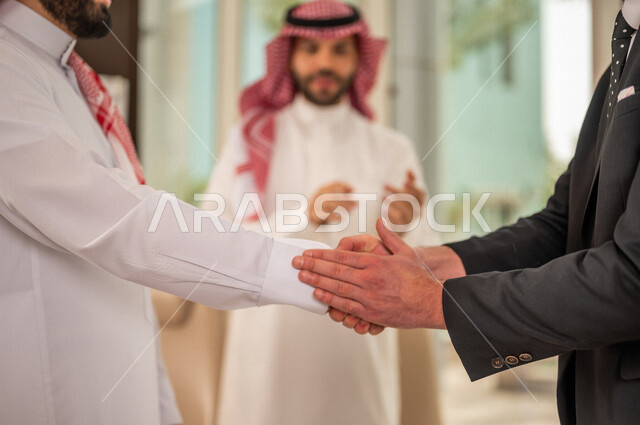 رجل اعمال عربي سعودي خليجي بالثوب السعودي التقليدي يتصافح مع عميل جديد يرتدي البدلة الرسمية، توقيع اتفاقية عمل لمشروع جديد، اتفاق عمل بين رجال الاعمال، صفقة ناجحة، تعاون تجاري، الوحدة والعمل الجماعي، الموافقة على شروط العمل، توقيع عقود العمل  