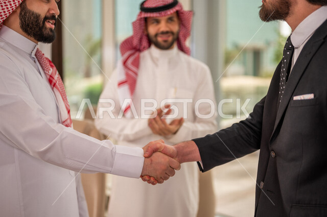 رجل اعمال عربي سعودي خليجي بالثوب السعودي التقليدي يتصافح مع عميل جديد يرتدي البدلة الرسمية، توقيع اتفاقية عمل لمشروع جديد، اتفاق عمل بين رجال الاعمال، صفقة ناجحة، تعاون تجاري، الوحدة والعمل الجماعي، الموافقة على شروط العمل، توقيع عقود العمل  
