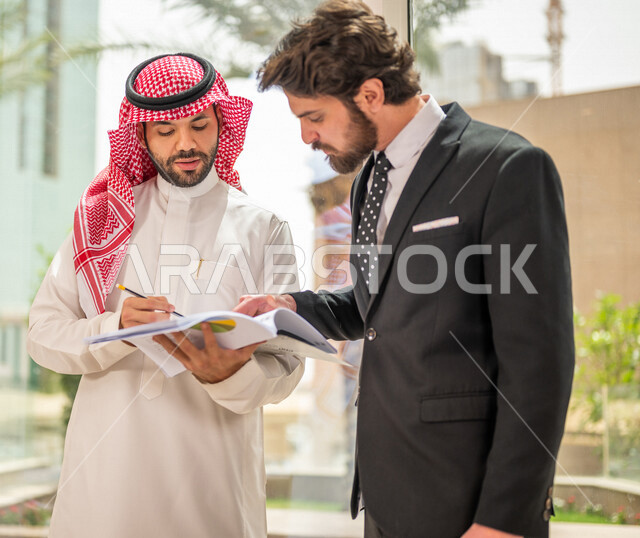A Saudi Arabian Gulf businessman in the traditional Saudi dress discusses with a client wearing a formal suit, a work agreement for a new project, analysis and planning of projects, work development and problem solving, unity and teamwork, achieving the c