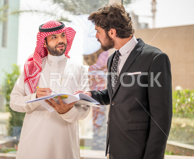 رجل اعمال عربي سعودي خليجي بالثوب السعودي التقليدي يتناقش مع عميل يرتدي البدلة الرسمية، اتفاقية عمل لمشروع جديد، تحليل وتخطيط المشاريع، تطوير العمل وحل المشكلات، الوحدة والعمل الجماعي، تحقيق اهداف الشركه