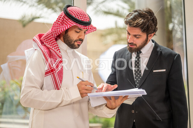A Saudi Arabian Gulf businessman in the traditional Saudi dress discusses with a client wearing a formal suit, a work agreement for a new project, analysis and planning of projects, work development and problem solving, unity and teamwork, achieving the c