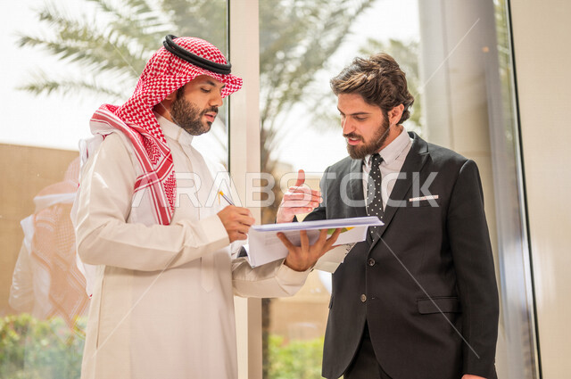 رجل اعمال عربي سعودي خليجي بالثوب السعودي التقليدي يتناقش مع عميل يرتدي البدلة الرسمية، اتفاقية عمل لمشروع جديد، تحليل وتخطيط المشاريع، تطوير العمل وحل المشكلات، الوحدة والعمل الجماعي، تحقيق اهداف الشركه
