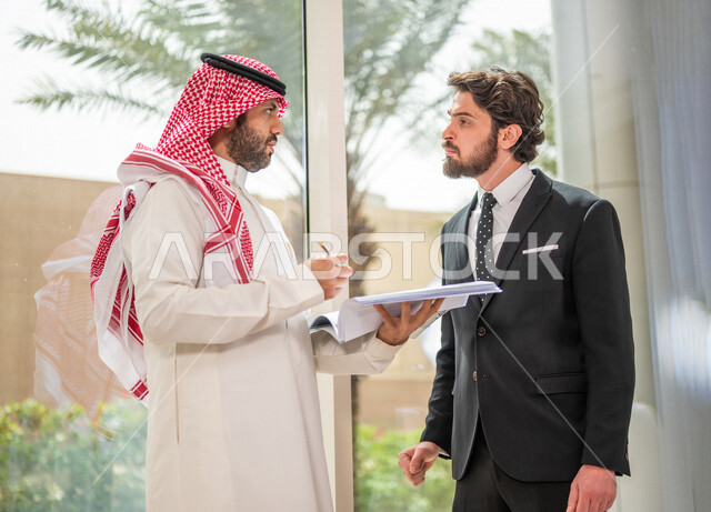 A Saudi Arabian Gulf businessman in the traditional Saudi dress discusses with a client wearing a formal suit, a work agreement for a new project, analysis and planning of projects, work development and problem solving, unity and teamwork, achieving the c