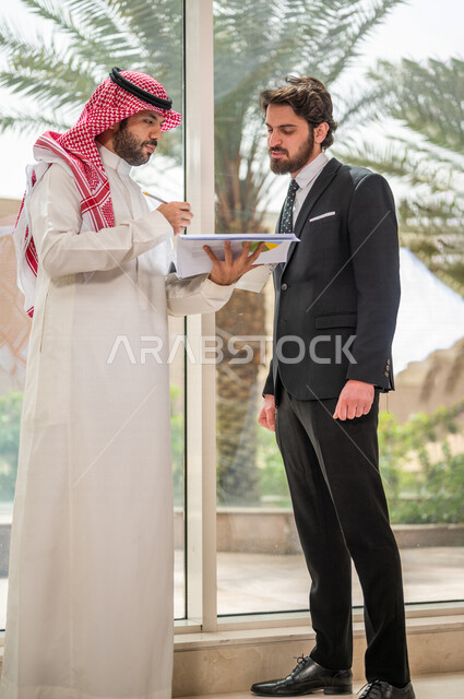رجل اعمال عربي سعودي خليجي بالثوب السعودي التقليدي يتناقش مع عميل يرتدي البدلة الرسمية، اتفاقية عمل لمشروع جديد، تحليل وتخطيط المشاريع، تطوير العمل وحل المشكلات، الوحدة والعمل الجماعي، تحقيق اهداف الشركه