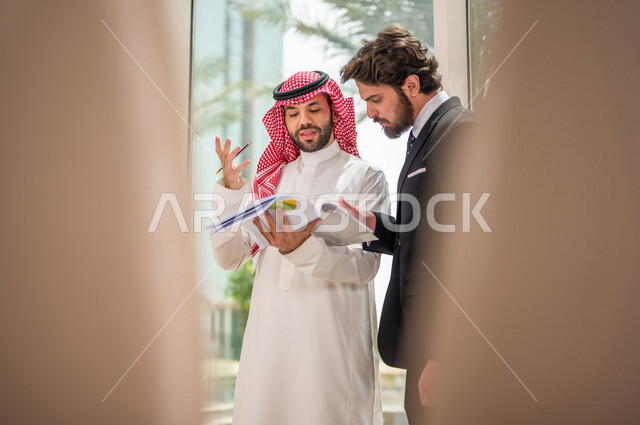 A Saudi Arabian Gulf businessman in the traditional Saudi dress discusses with a client wearing a formal suit, a work agreement for a new project, analysis and planning of projects, work development and problem solving, unity and teamwork, achieving the c