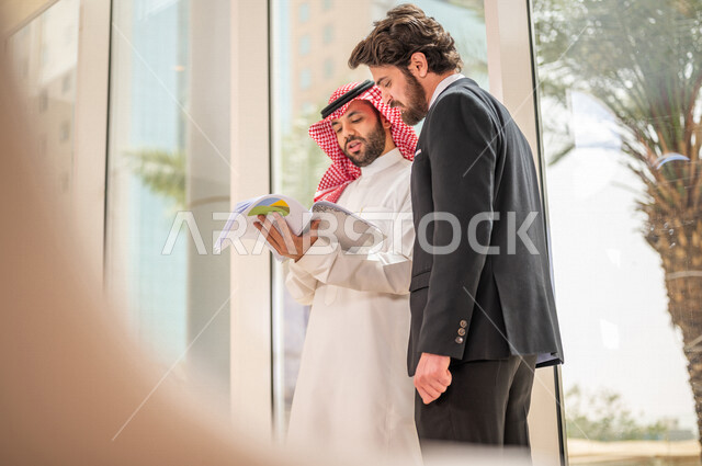 رجل اعمال عربي سعودي خليجي بالثوب السعودي التقليدي يتناقش مع عميل يرتدي البدلة الرسمية، اتفاقية عمل لمشروع جديد، تحليل وتخطيط المشاريع، تطوير العمل وحل المشكلات، الوحدة والعمل الجماعي، تحقيق اهداف الشركه