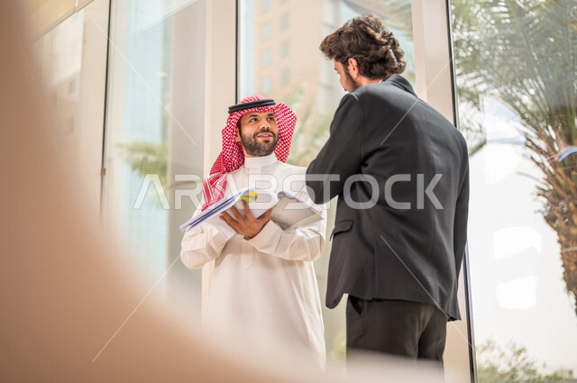 رجل اعمال عربي سعودي خليجي بالثوب السعودي التقليدي يتناقش مع عميل يرتدي البدلة الرسمية، اتفاقية عمل لمشروع جديد، تحليل وتخطيط المشاريع، تطوير العمل وحل المشكلات، الوحدة والعمل الجماعي، تحقيق اهداف الشركه