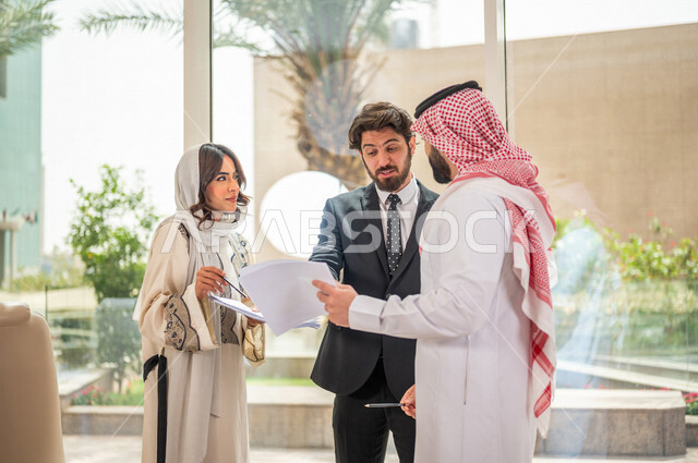 رجل اعمال عربي سعودي خليجي بالثوب السعودي التقليدي يتناقش مع عميل يرتدي البدلة الرسمية، اتفاقية عمل لمشروع جديد، سيدة اعمال عربية سعودية خليجية تعمل على تحليل وتخطيط المشاريع، تطوير العمل وحل المشكلات، الوحدة والعمل الجماعي، تحقيق اهداف الشركه 
