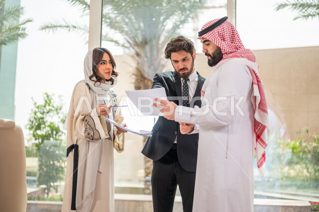 رجل اعمال عربي سعودي خليجي بالثوب السعودي التقليدي يتناقش مع عميل يرتدي البدلة الرسمية، اتفاقية عمل لمشروع جديد، سيدة اعمال عربية سعودية خليجية تعمل على تحليل وتخطيط المشاريع، تطوير العمل وحل المشكلات، الوحدة والعمل الجماعي، تحقيق اهداف الشركه 