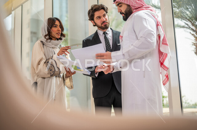 رجل اعمال عربي سعودي خليجي بالثوب السعودي التقليدي يتناقش مع عميل يرتدي البدلة الرسمية، اتفاقية عمل لمشروع جديد، سيدة اعمال عربية سعودية خليجية تعمل على تحليل وتخطيط المشاريع، تطوير العمل وحل المشكلات، الوحدة والعمل الجماعي، تحقيق اهداف الشركه 