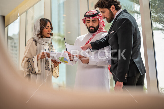 A Saudi Arabian Gulf businessman in the traditional Saudi dress discusses with a client wearing a formal suit, a work agreement for a new project, a Saudi Arabian Gulf business woman working on project analysis and planning, work development and problem s