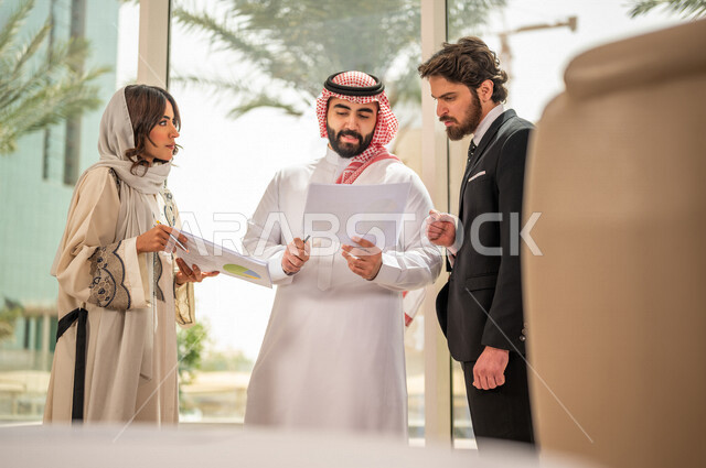 رجل اعمال عربي سعودي خليجي بالثوب السعودي التقليدي يتناقش مع عميل يرتدي البدلة الرسمية، اتفاقية عمل لمشروع جديد، سيدة اعمال عربية سعودية خليجية تعمل على تحليل وتخطيط المشاريع، تطوير العمل وحل المشكلات، الوحدة والعمل الجماعي، تحقيق اهداف الشركه 