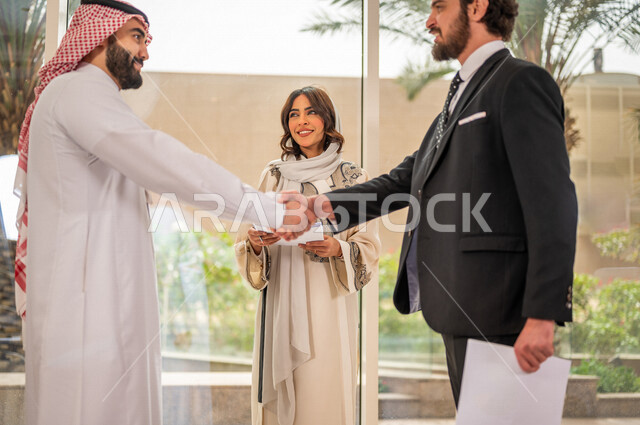رجل اعمال عربي سعودي خليجي بالثوب السعودي التقليدي يتصافح مع عميل جديد يرتدي البدلة الرسمية، سيدة اعمال خليجية تشارك في نجاح صفقات العمل، توقيع اتفاقية عمل لمشروع جديد، صفقة ناجحة، الوحدة والعمل الجماعي، الموافقة على شروط العمل، توقيع عقود العمل