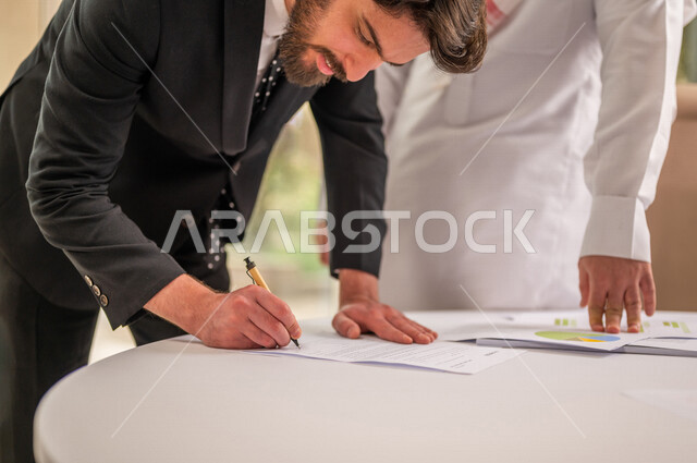 رجل اعمال عربي سعودي خليجي بالثوب السعودي التقليدي يقوم بتوقيع اتفاقية عمل لمشروع جديد مع عميل يرتدي البدلة الرسمية، صفقة ناجحة، الوحدة والعمل الجماعي، التحقق والتدقيق في شروط العقد، قراءة البنود الخاصة بالعقد، الموافقة على الشروط، التوقيع على العقود 