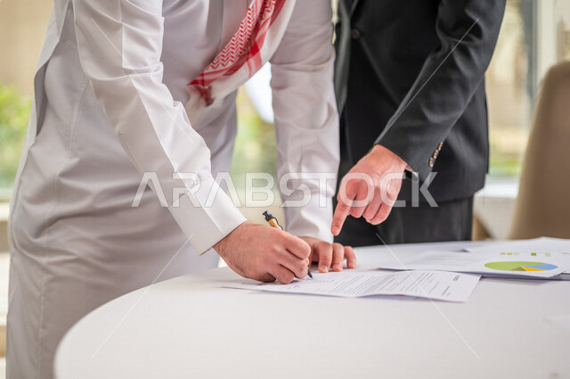 رجل اعمال عربي سعودي خليجي بالثوب السعودي التقليدي يقوم بتوقيع اتفاقية عمل لمشروع جديد مع عميل يرتدي البدلة الرسمية، صفقة ناجحة، الوحدة والعمل الجماعي، التحقق والتدقيق في شروط العقد، قراءة البنود الخاصة بالعقد، الموافقة على الشروط، التوقيع على العقود 