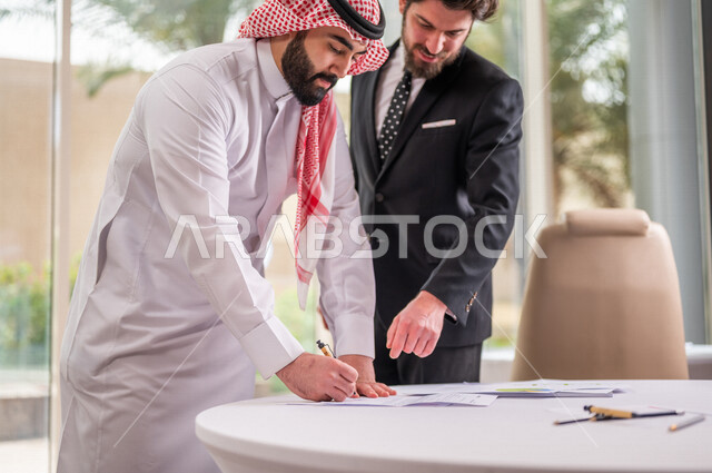رجل اعمال عربي سعودي خليجي بالثوب السعودي التقليدي يقوم بتوقيع اتفاقية عمل لمشروع جديد مع عميل يرتدي البدلة الرسمية، صفقة ناجحة، الوحدة والعمل الجماعي، التحقق والتدقيق في شروط العقد، قراءة البنود الخاصة بالعقد، الموافقة على الشروط، التوقيع على العقود 