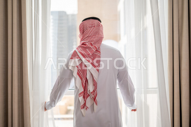 رجل عربي خليجي سعودي يقف امام النافذة، غرفة فندقية، النظر الى الخارج من خلال النافذة، إشراقة يوم جديد، مفهوم الهمة والنشاط، التخخطيط المستقبلي، الرؤية المستقبلية، سياحة وفندقة