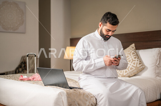 رجل عربي خليجي سعودي يجلس في غرفة فندقية ويستخدم الهاتف المحمول، إنجاز المهام من خلال الحاسوب المحمول، إستخدام تقنيات حديثة، العمل عن بعد، مهن ووظائف، تخطيط العمل، مشروع عمل جديد،  تنظيم جدول الاعمال، مفهوم إدارة الأعمال، رحلة عمل، سياحة وفندقة