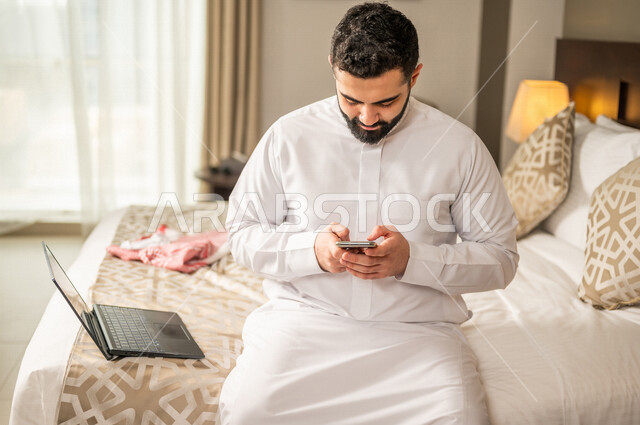 رجل عربي خليجي سعودي يجلس في غرفة فندقية ويستخدم الهاتف المحمول، إنجاز المهام من خلال الحاسوب المحمول، إستخدام تقنيات حديثة، العمل عن بعد، مهن ووظائف، تخطيط العمل، مشروع عمل جديد،  تنظيم جدول الاعمال، مفهوم إدارة الأعمال، رحلة عمل، سياحة وفندقة