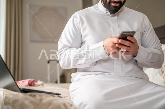 رجل عربي خليجي سعودي يجلس في غرفة فندقية ويستخدم الهاتف المحمول، إنجاز المهام من خلال الحاسوب المحمول، إستخدام تقنيات حديثة، العمل عن بعد، مهن ووظائف، تخطيط العمل، مشروع عمل جديد،  تنظيم جدول الاعمال، مفهوم إدارة الأعمال، رحلة عمل، سياحة وفندقة