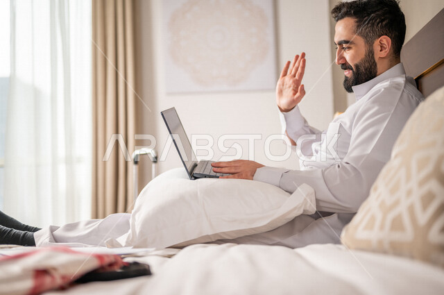 رجل عربي خليجي سعودي يقوم باجراءة مكالمة فيديو من خلال الحاسوب المحمول، اجتماع عمل عن بعد، القاء التحية من خلال شاشة اللابتوب، تقنيات حديثة، دردشة وتواصل، علاقات اجتماعية، سياحة وفندقة