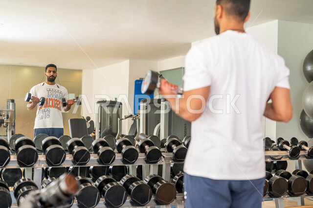 رجل عربي خليجي سعودي يرتدي الملابس الرياضية ويقوم بالتمارين الرياضية باستخدام الدمبل ، تمارين بناء العضلات وتقويتها ، ممارسة تمارين رفع الاوزان، نادي الألعاب الرياضية، الصحة واللياقة البدنية