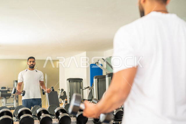 رجل عربي خليجي سعودي يرتدي الملابس الرياضية ويقوم بالتمارين الرياضية باستخدام الدمبل ، تمارين بناء العضلات وتقويتها ، ممارسة تمارين رفع الاوزان، نادي الألعاب الرياضية، الصحة واللياقة البدنية