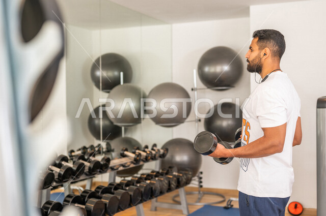 رجل عربي خليجي سعودي يرتدي الملابس الرياضية ويقوم بالتمارين الرياضية باستخدام الدمبل ، تمارين بناء العضلات وتقويتها ، ممارسة تمارين رفع الاوزان، نادي الألعاب الرياضية، الصحة واللياقة البدنية