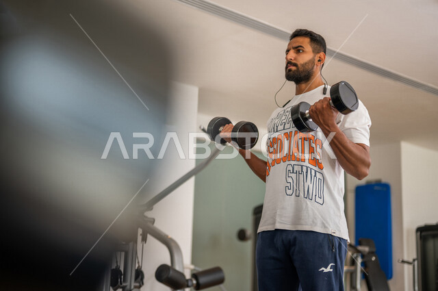 رجل عربي خليجي سعودي يرتدي الملابس الرياضية ويقوم بالتمارين الرياضية باستخدام الدمبل ، تمارين بناء العضلات وتقويتها ، ممارسة تمارين رفع الاوزان، نادي الألعاب الرياضية، الصحة واللياقة البدنية