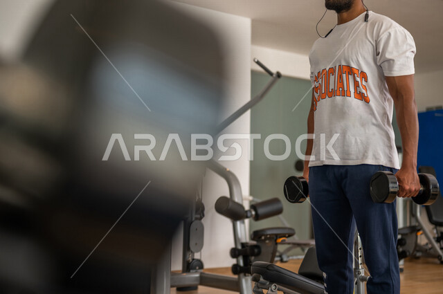 رجل عربي خليجي سعودي يرتدي الملابس الرياضية ويقوم بالتمارين الرياضية باستخدام الدمبل ، تمارين بناء العضلات وتقويتها ، ممارسة تمارين رفع الاوزان، نادي الألعاب الرياضية، الصحة واللياقة البدنية