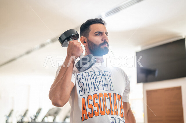 رجل عربي خليجي سعودي يرتدي الملابس الرياضية ويقوم بالتمارين الرياضية باستخدام الدمبل ، تمارين بناء العضلات وتقويتها ، ممارسة تمارين رفع الاوزان، نادي الألعاب الرياضية، الصحة واللياقة البدنية