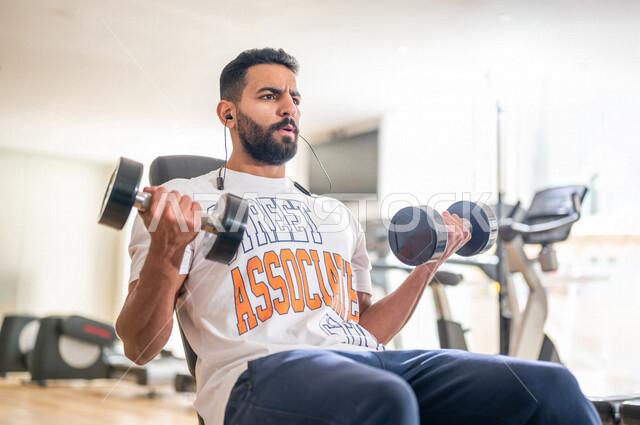 رجل عربي خليجي سعودي يرتدي الملابس الرياضية ويقوم بالتمارين الرياضية باستخدام الدمبل ، تمارين بناء العضلات وتقويتها ، ممارسة تمارين رفع الاوزان، نادي الألعاب الرياضية، الصحة واللياقة البدنية