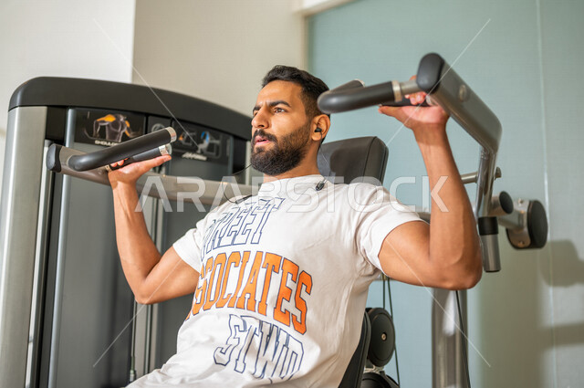 رجل عربي خليجي سعودي يرتدي الملابس الرياضية ويقوم بالتمارين الرياضية باستخدام جهاز الفراشة لتقوية العضلات، تمارين بناء العضلات وتقويتها ، ممارسة تمارين رفع الاوزان، نادي الألعاب الرياضية، الصحة واللياقة البدنية