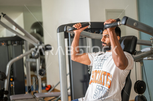 رجل عربي خليجي سعودي يرتدي الملابس الرياضية ويقوم بالتمارين الرياضية باستخدام جهاز الفراشة لتقوية العضلات، تمارين بناء العضلات وتقويتها ، ممارسة تمارين رفع الاوزان، نادي الألعاب الرياضية، الصحة واللياقة البدنية