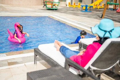 A Saudi Arabian Gulf woman wearing a swimming suit and enjoying sitting on the deck chair, two Saudi Arabian Gulf children enjoying swimming in the water pool, relaxation and entertainment, a leisure tourist trip, a happy summer vacation