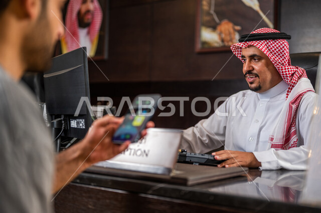 سائح عربي خليجي سعودي يستخدم الهاتف المحمول أمام موظف إستقبال خليجي سعودي داخل الفندق ، الدفع و تأكيد الحجوزات من خلال تطبيق الهاتف الذكي ، إجراءات الحجز و الدخول ، خدمة العملاء في الفندق السياحي ، سياحة و فندقة