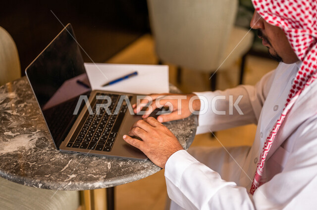 صورة مقربة لرجل أعمال عربي خليجي سعودي يستخدم الحاسوب المحمول يجلس في أحد الفنادق الخليجية ، انجاز مهام العمل ، تحقيق أهداف الشركة و العمل ، متابعة وتطوير الأعمال ، العمل عن بعد ، كتابة ملاحظات و تقييم الأعمال و المشاريع