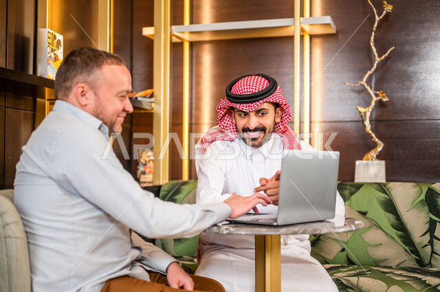 رجلان أعمال عربيان خليجيان سعوديان يستخدمان الحاسوب المحمول و يتناقشان بالعمل ، شركاء عمل يجتمعان في أحد الفنادق الخليجية ، إتفاق عمل ، الشراكة على الأعمال و المشاريع التجارية ، صفقات عمل و تعاون بين الشركات  