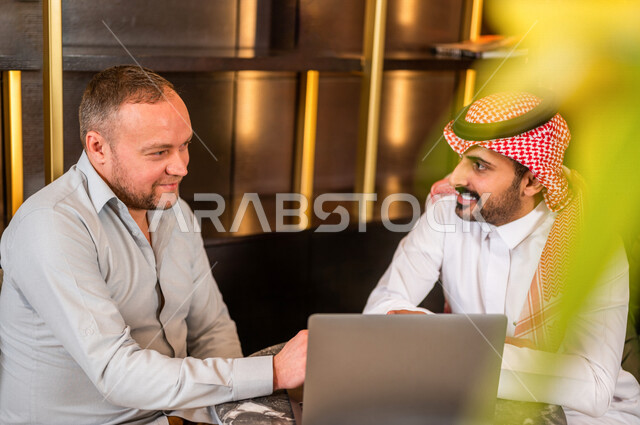 رجلان أعمال عربيان خليجيان سعوديان يستخدمان الحاسوب المحمول و يتناقشان بالعمل ، شركاء عمل يجتمعان في أحد الفنادق الخليجية ، إتفاق عمل ، الشراكة على الأعمال و المشاريع التجارية ، صفقات عمل و تعاون بين الشركات  