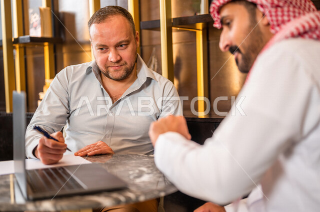 رجلان أعمال عربيان خليجيان سعوديان يستخدمان الحاسوب المحمول و يتناقشان بالعمل ، شركاء عمل يجتمعان في أحد الفنادق الخليجية ، إتفاق عمل ، الشراكة على الأعمال و المشاريع التجارية ، صفقات عمل و تعاون بين الشركات  