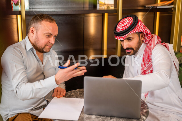 رجلان أعمال عربيان خليجيان سعوديان يستخدمان الحاسوب المحمول و يتناقشان بالعمل ، شركاء عمل يجتمعان في أحد الفنادق الخليجية ، إتفاق عمل ، الشراكة على الأعمال و المشاريع التجارية ، صفقات عمل و تعاون بين الشركات  