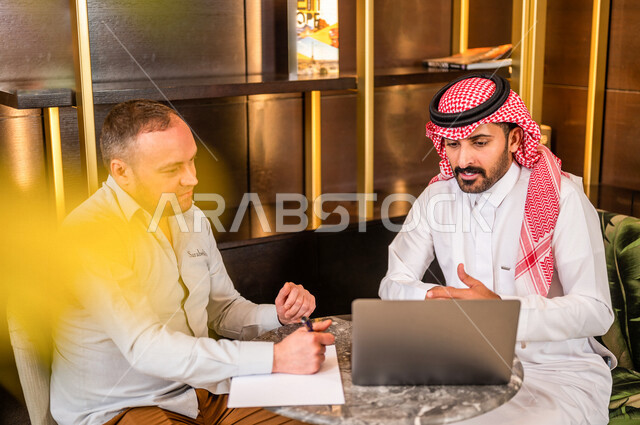 رجلان أعمال عربيان خليجيان سعوديان يستخدمان الحاسوب المحمول و يتناقشان بالعمل ، شركاء عمل يجتمعان في أحد الفنادق الخليجية ، إتفاق عمل ، الشراكة على الأعمال و المشاريع التجارية ، صفقات عمل و تعاون بين الشركات  